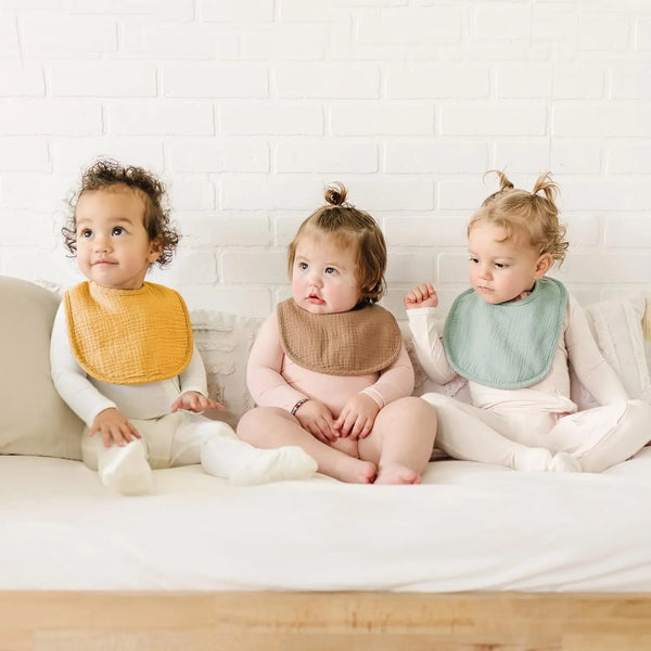 Tres bebés con pañuelos de muselina amarillos y verdes sentados en una cama blanca