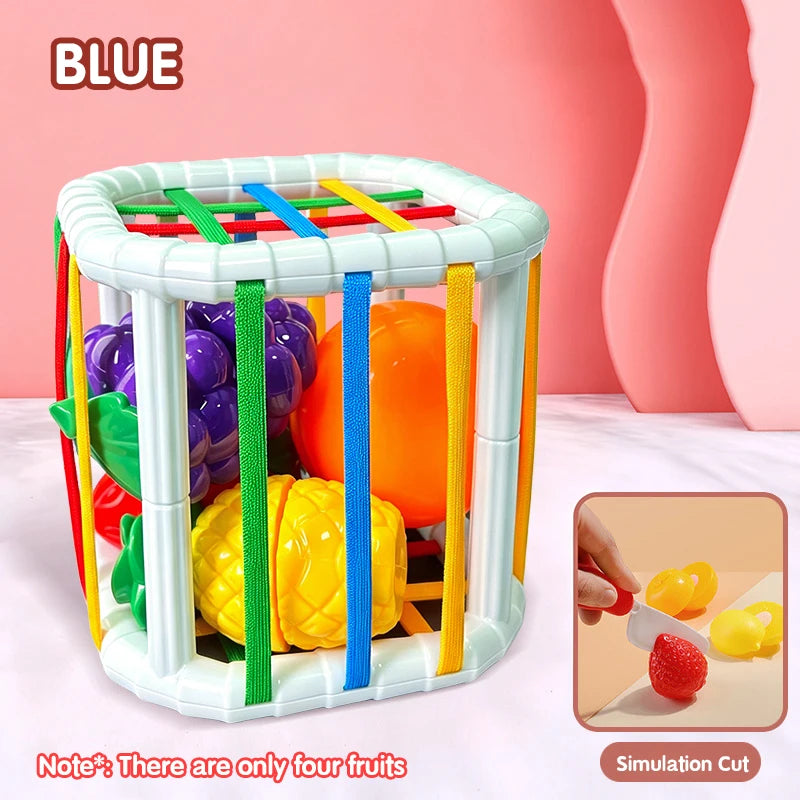 A white plastic toy basket with colored vertical bars holding four different fruits: purple grapes, green apples, orange, and