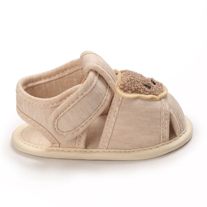 A pair of beige soft sole sandals for a baby, featuring a bear-shaped detail.