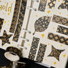A close-up of a notebook decorated with black and gold foil patterned paper and several rolls of matching washi tape.