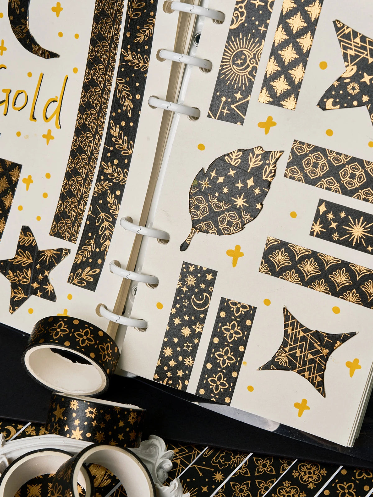 A close-up of a notebook decorated with black and gold foil patterned paper and several rolls of matching washi tape.