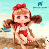A small, rosy-cheeked doll with reddish hair is seated in a wooden deck chair outdoors.