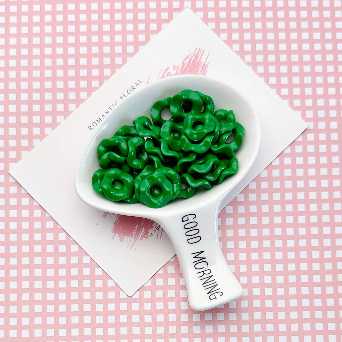 A white spoon holds a pile of bright green floral acrylic beads on a pink checkered background.