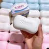 A hand holds a white soap ball squishy fidget toy surrounded by other pink and blue soap products.