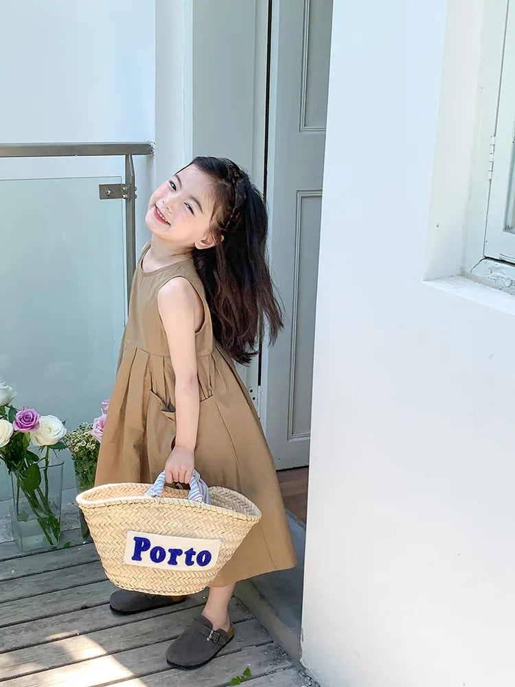A young girl wearing a beige sleeveless cotton sundress stands outdoors near a white door.