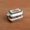 A miniature doll picnic lunch box with a lid featuring a floral design, shown on a wooden surface.