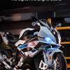 A white and black 1:12 scale die-cast model motorcycle is shown with its rear wheel triggers pressed, featuring bright lights