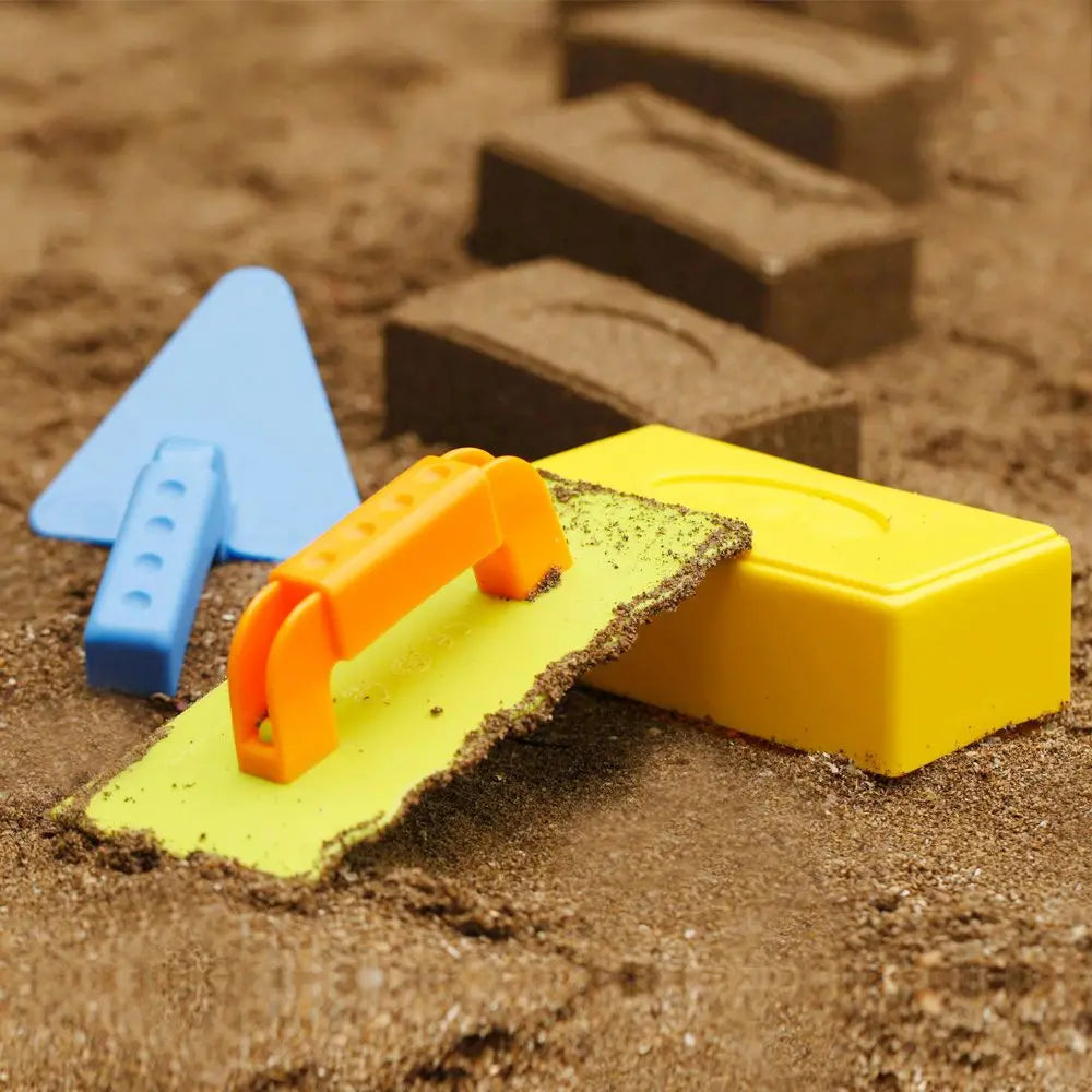 A yellow sand brick mold is shown next to blue and brown plastic pieces on a sandy surface.