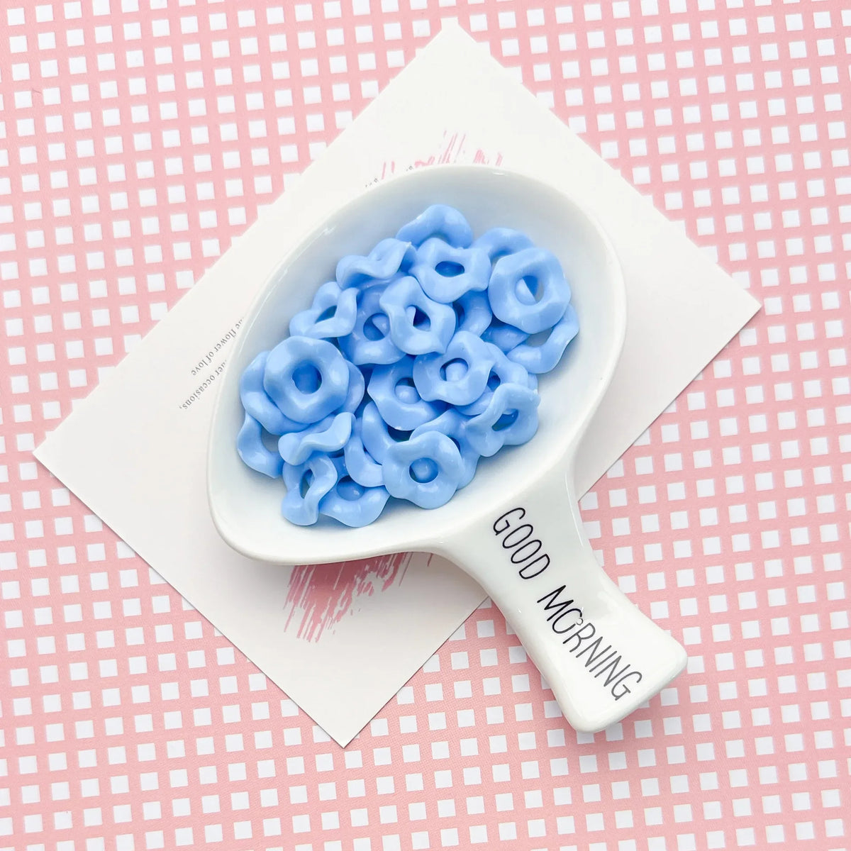 A white spoon holds a cluster of light blue acrylic beads with a floral edge on a pink patterned background.