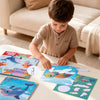 A child is assembling a sticker art puzzle kit featuring dinosaur and animal designs on a white table.