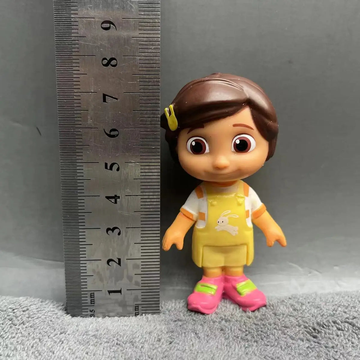 A playful cartoon PVC figurine of a girl with brown hair and yellow clothing standing next to a ruler.