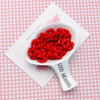 A white spoon holds bright red, textured acrylic beads with a floral edge on a pink patterned background.