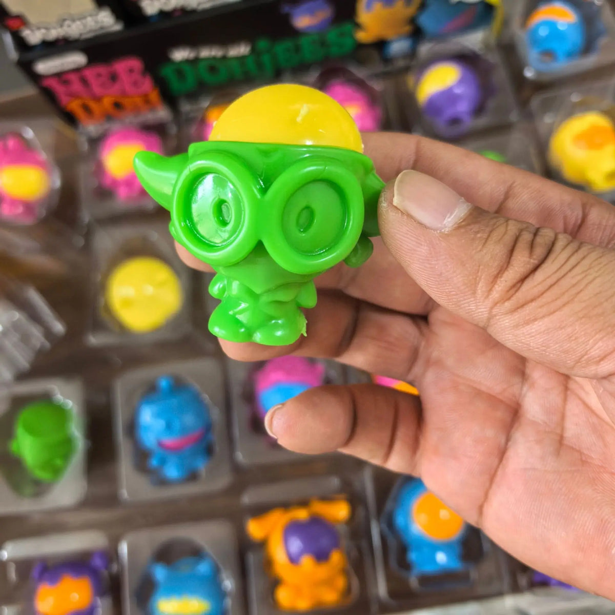 A hand holds a bright green squishy fidget toy with yellow eyes, displayed in a clear plastic container.