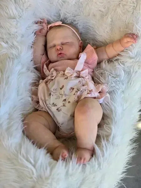 A newborn baby doll with hand-painted skin is lying peacefully in a fluffy white fur blanket.