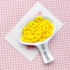 A white spoon holds bright yellow acrylic beads with a floral edge on a pink checkered background.