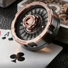 A close-up shot of a polished rose gold roulette fidget spinner resting on a dark surface next to playing cards and dice.