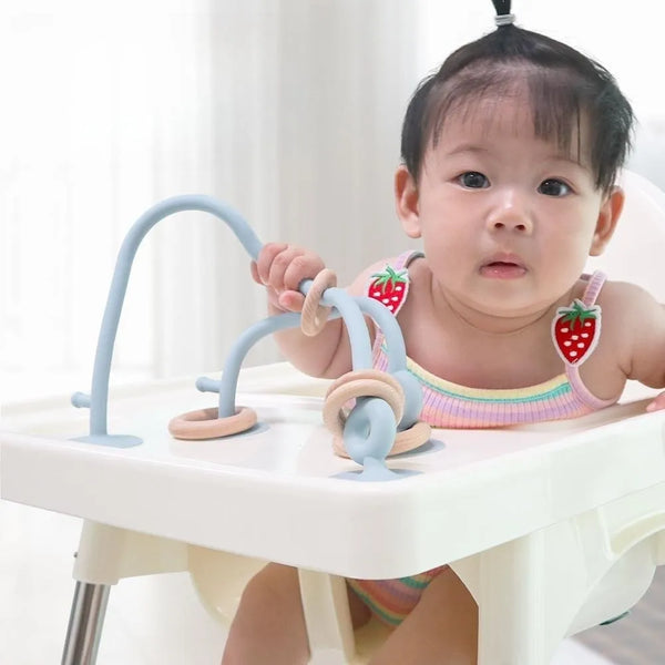 A baby is using a soft silicone teether ring on a white feeding tray.