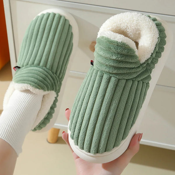 A pair of green faux fur slippers with white trim and sole being held by a hand.