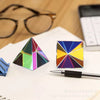 A geometric prism cube displaying various vibrant colors, resting on a white surface next to a pen and eyeglasses.