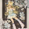 A collectible figure of a blonde girl with light blue hair and a white dress, displayed in a wooden frame surrounded by lace 