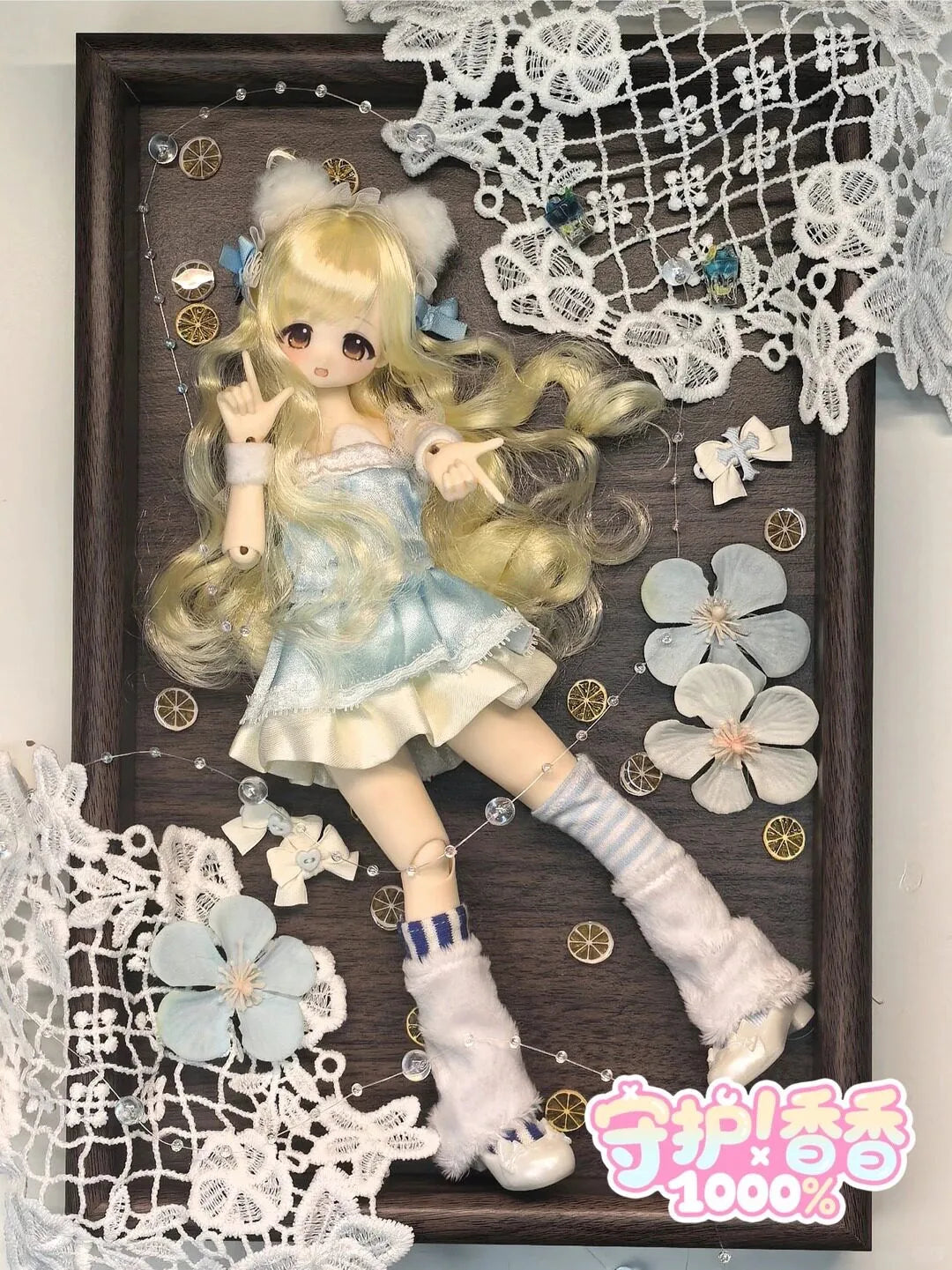 A collectible figure of a blonde girl with light blue hair and a white dress, displayed in a wooden frame surrounded by lace 