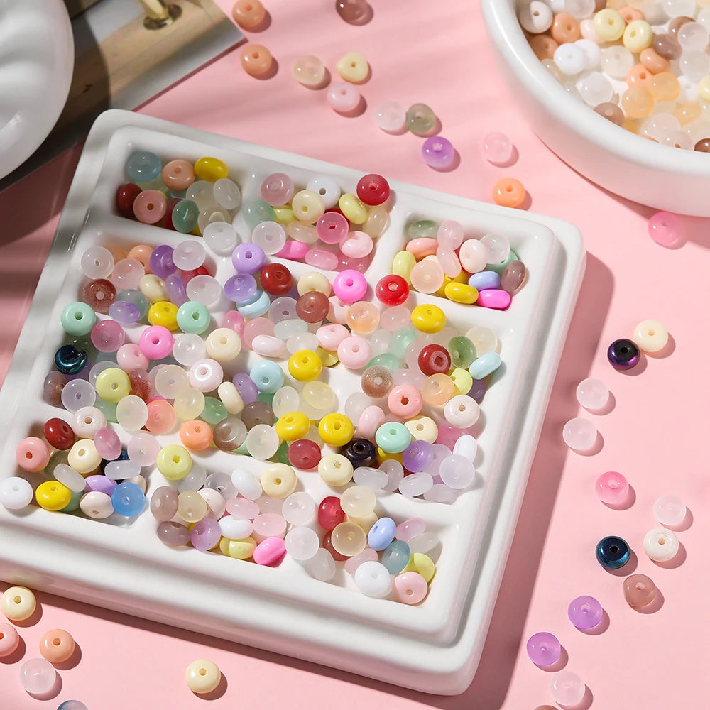 A white tray filled with various colored glass spacer beads, including jade tones, scattered on a pink surface.