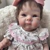 A lifelike 18-inch reborn baby doll with hand-painted details wearing a white floral dress and pink bow.