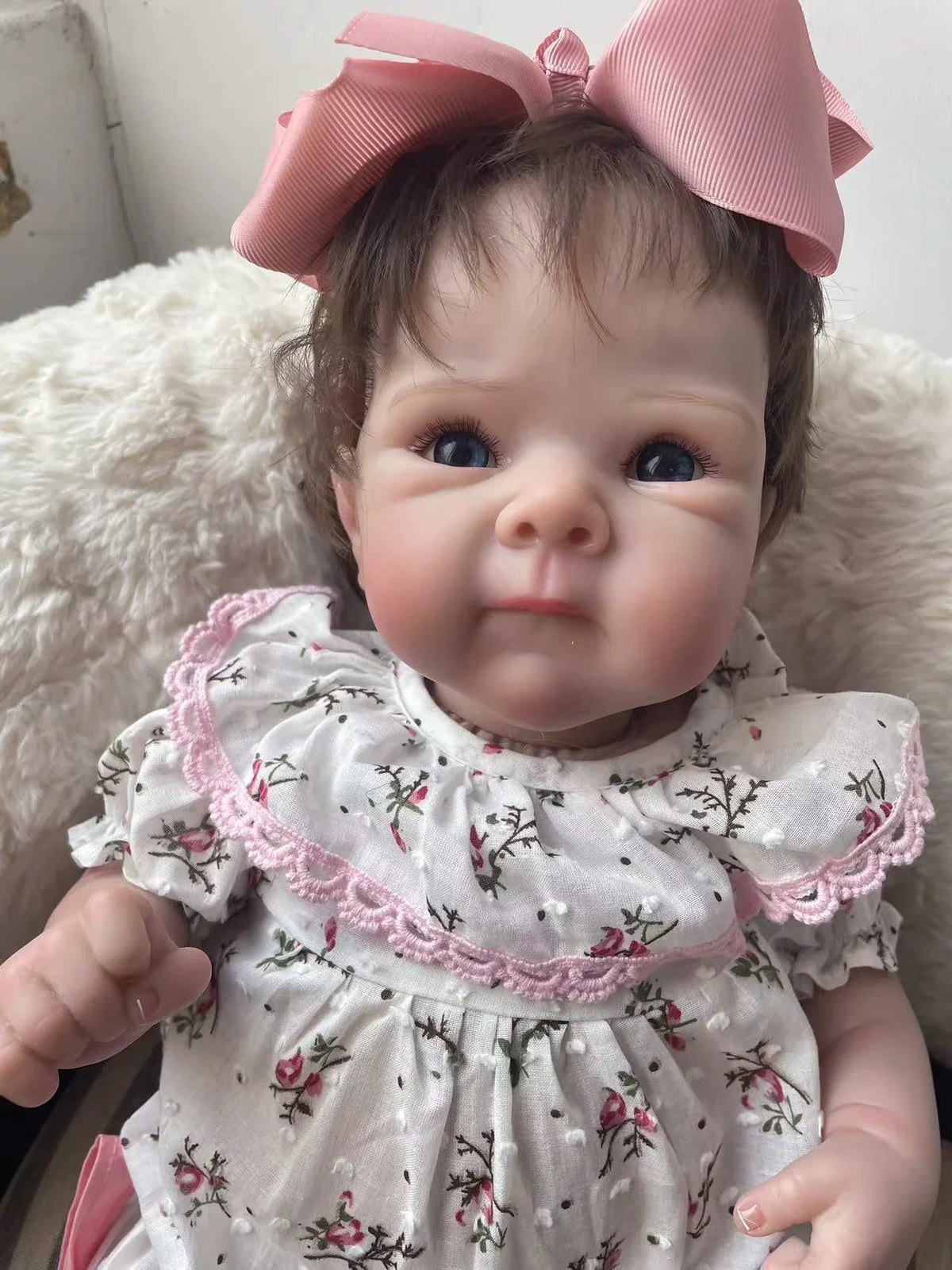 A lifelike 18-inch reborn baby doll with hand-painted details wearing a white floral dress and pink bow.