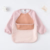 A pink waterproof long-sleeved baby bib with a beige insert and a white pom-pom nearby.