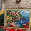 A large diamond painting depicting a coastal landscape with buildings, cliffs, and blue sea under a cloudy sky.