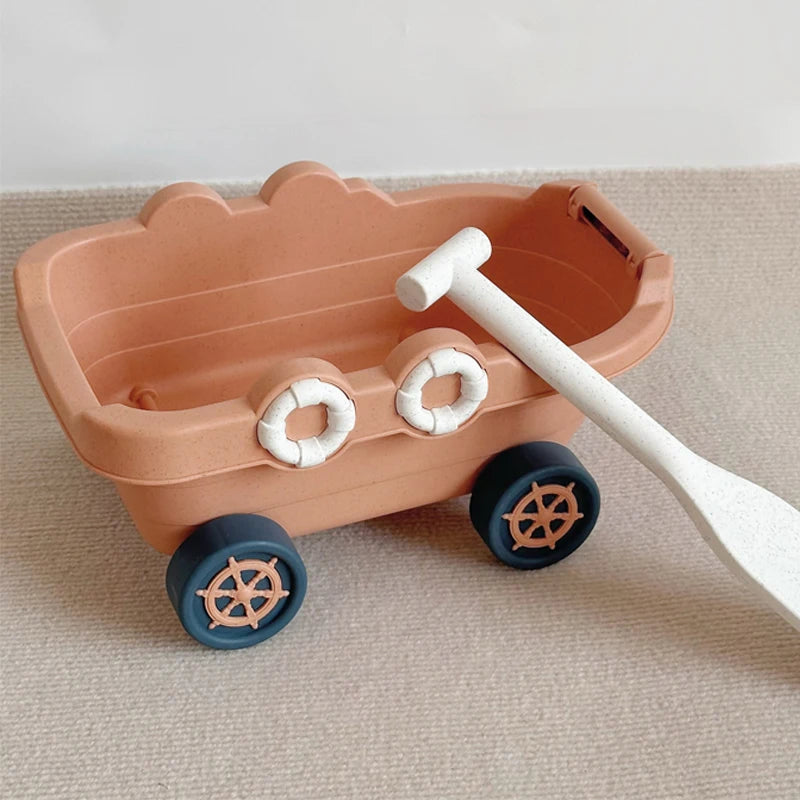 A pink kids sand and water play set featuring a pull-along bulldozer trolley with wooden wheels and a white shovel.