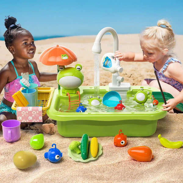 Two toddlers play with a green electric kitchen sink set filled with water and various colorful plastic food items on a sandy