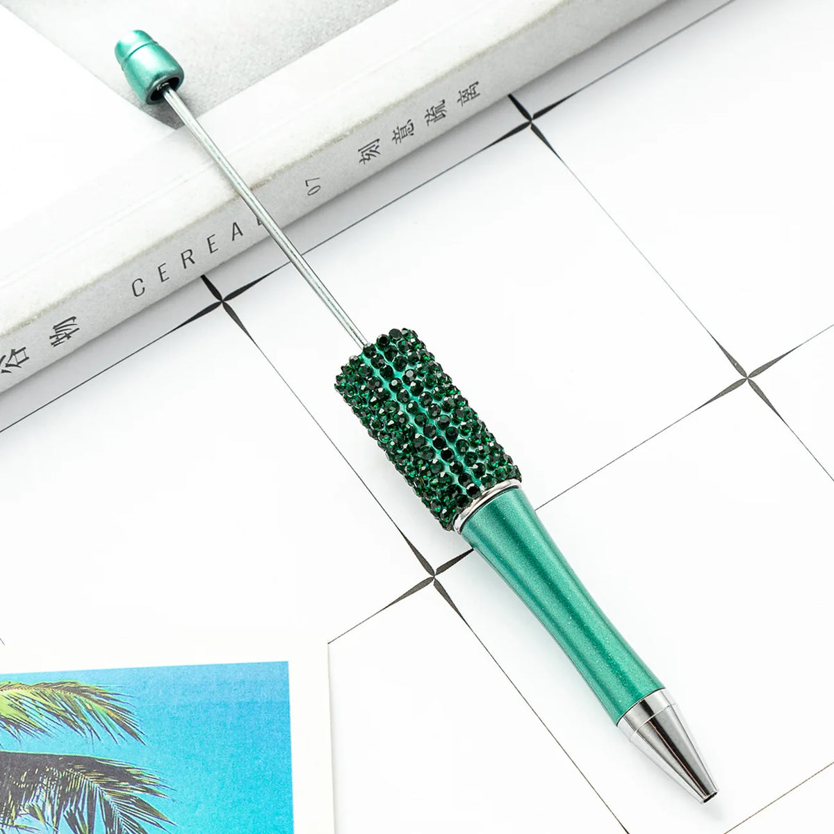 A green beaded ballpoint pen is shown resting on a white surface next to a book.