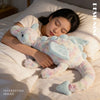 A light blue and pink rainbow dragon plush pillow is resting on a white sheet next to a person's head.