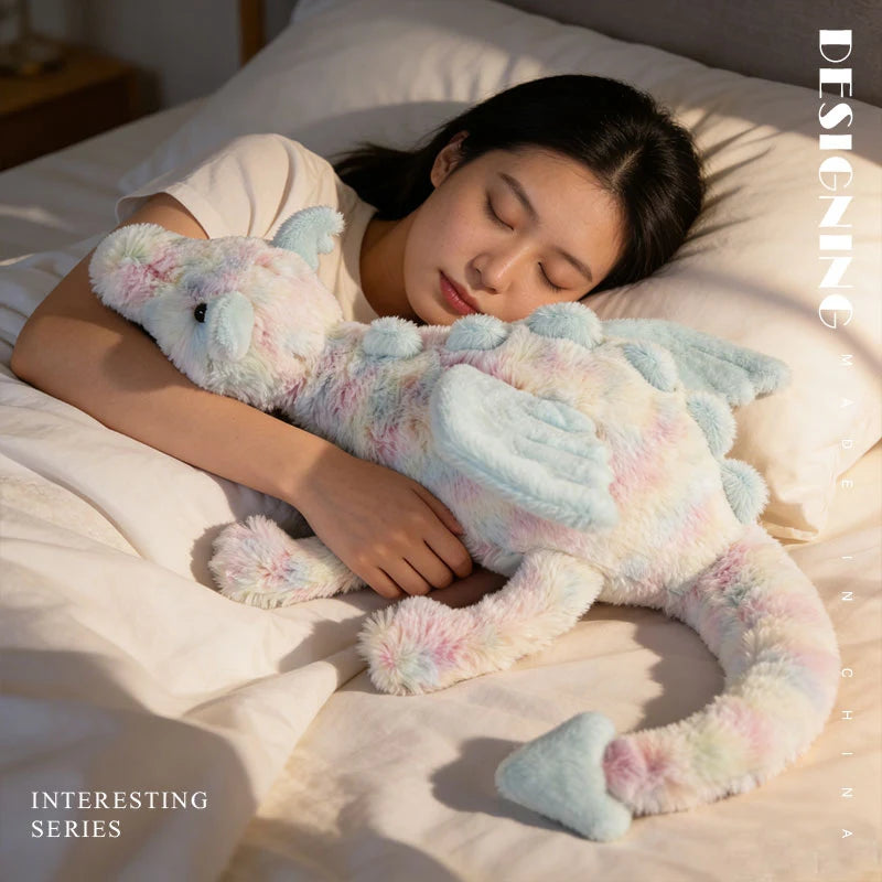 A light blue and pink rainbow dragon plush pillow is resting on a white sheet next to a person's head.