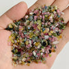 A hand holds a pile of natural tumbled stone chips in various shades of pink, green, and yellow.