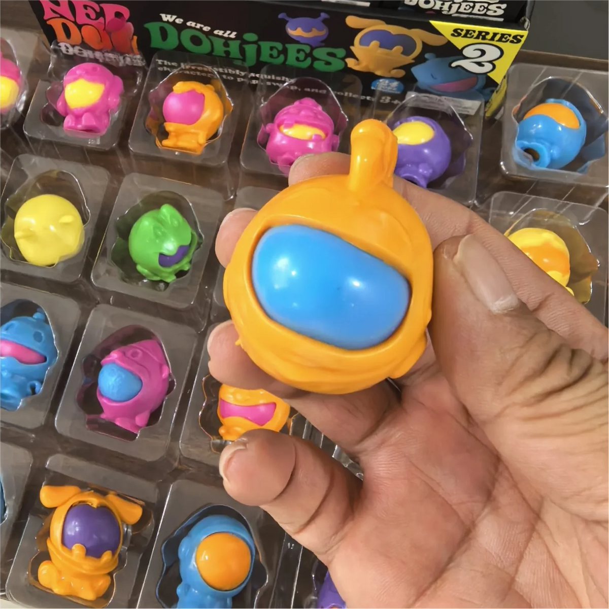 A hand holds an orange squishy fidget toy with a blue oval center, displayed in its packaging.