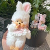 A white fluffy rabbit plush toy with pink ears is held against a background of green foliage and smaller similar toys.