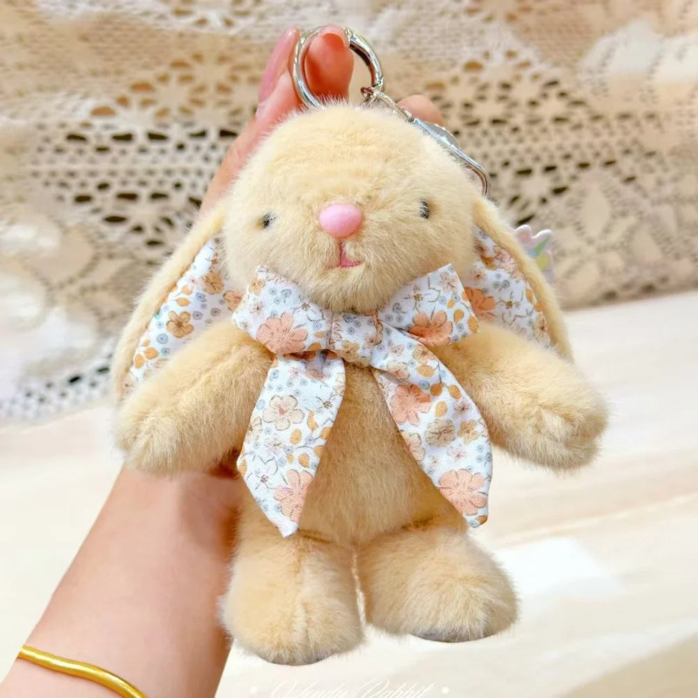 A fluffy light brown bunny plush toy with a pink nose and a floral bow charm being held by a hand.