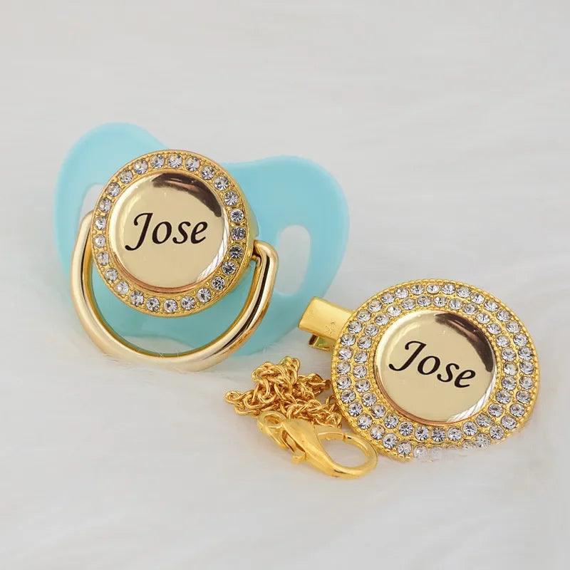 A gold-toned crystal pacifier and clip set featuring a circular charm engraved with the name Jose on a light blue silicone ba