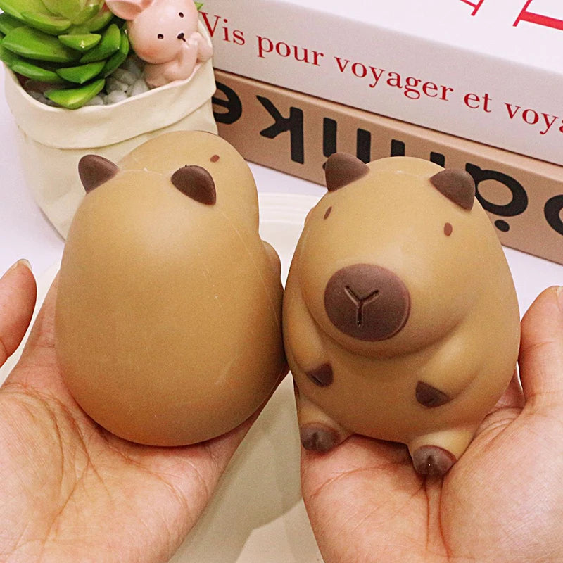 A pair of smooth, beige capybara squeeze stress relief toys are held in a hand next to a box.