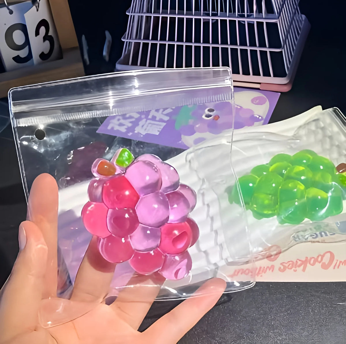 A hand holds a clear plastic bag containing pink and purple squishy grape-shaped fidget toys.