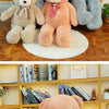Three large plush teddy bears in gray and orange sitting on a wooden floor, with a smaller bear resting on a bed.