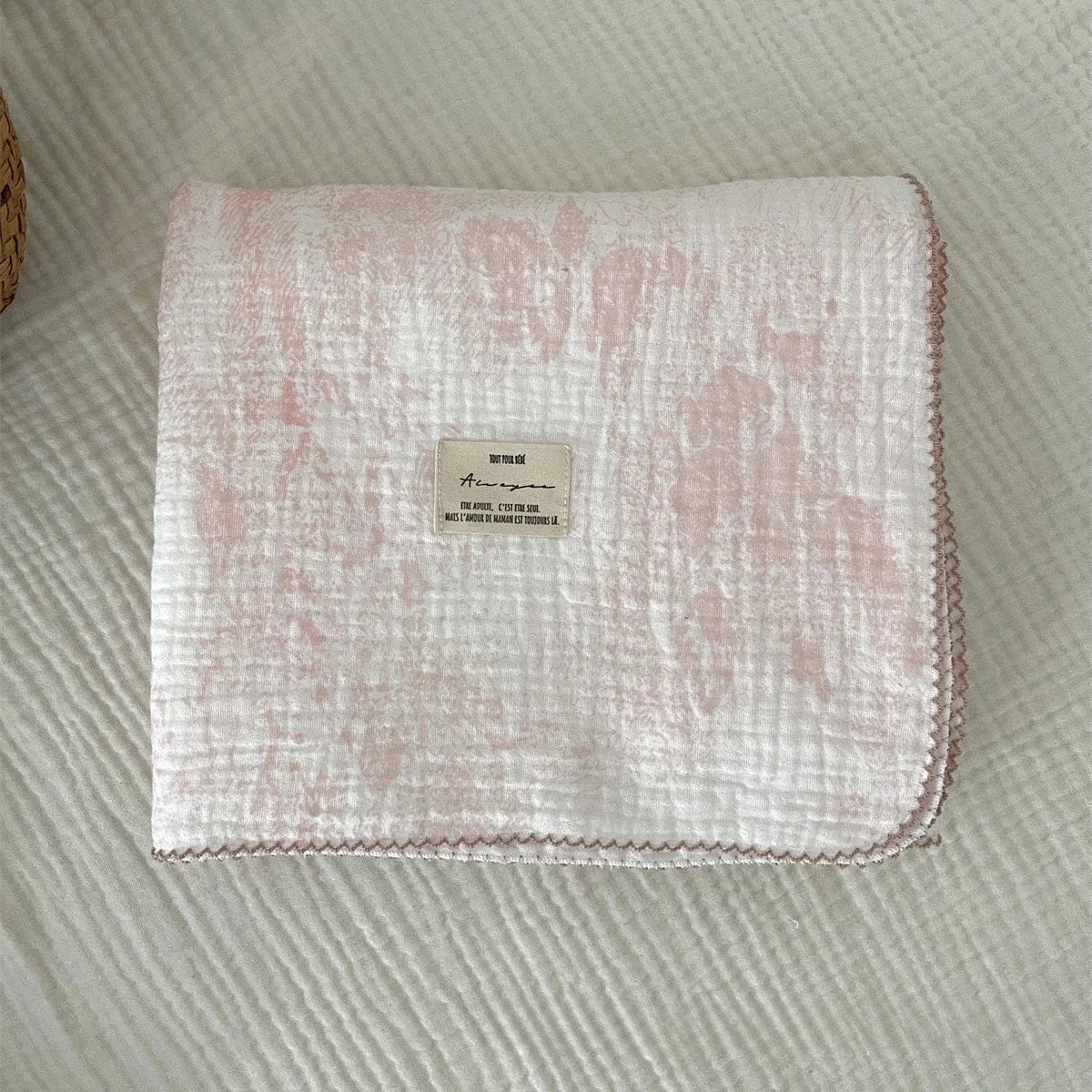 A pink muslin swaddle blanket with a floral print is displayed on a white textured surface.