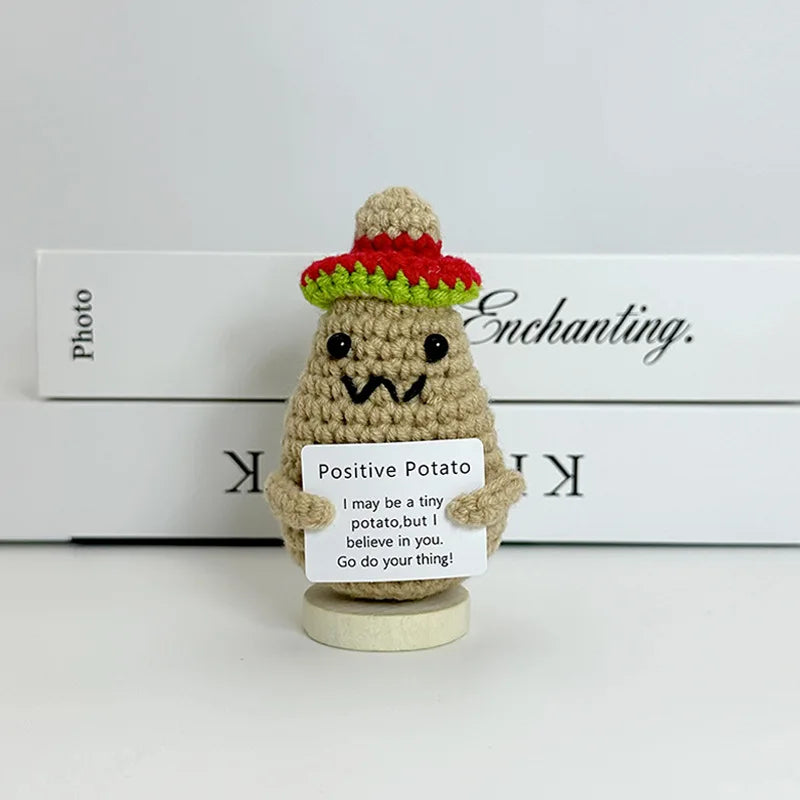 A handwoven crochet ornament shaped like a potato with a red hat and a sign encouraging positivity.