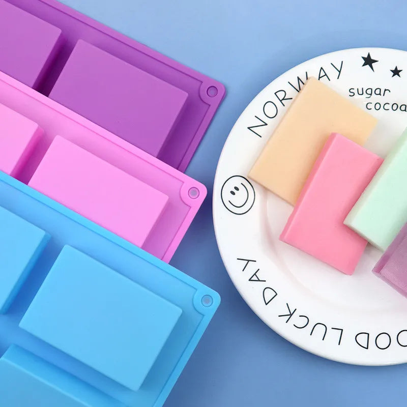 A white silicone soap mold with six square cavities is shown next to purple and pink molds, labeled for sugar and cocoa.