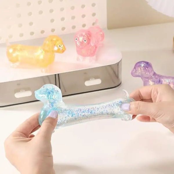 A hand holds a clear glitter dachshund squishy stress relief toy next to pink and purple plush toys.