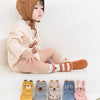 A baby wearing a beige outfit and a brown hood is sitting, with several pairs of cute cartoon animal non-slip socks laid out 
