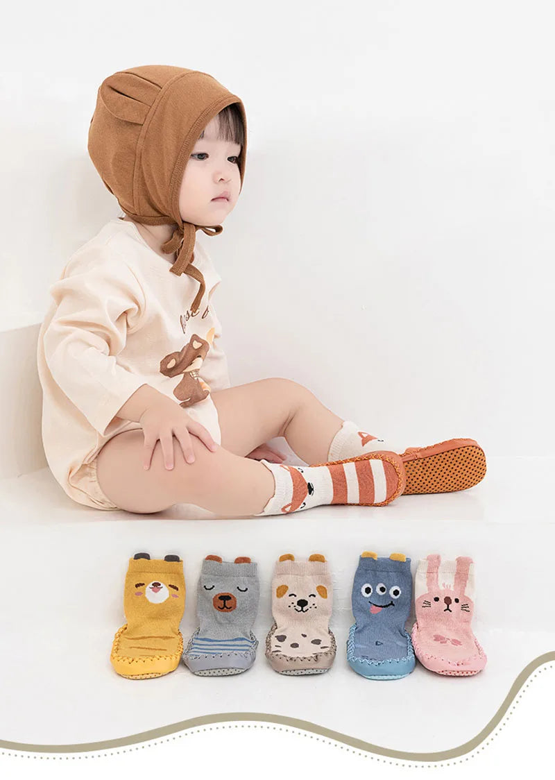 A baby wearing a beige outfit and a brown hood is sitting, with several pairs of cute cartoon animal non-slip socks laid out 