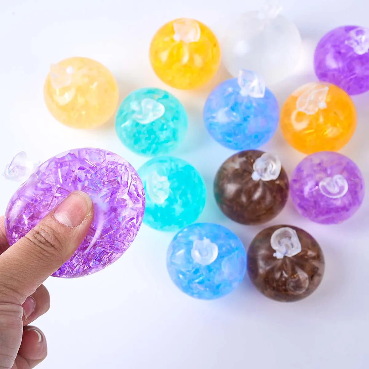 A hand holds a purple squeeze ball next to a set of six colorful, translucent stress relief balls.